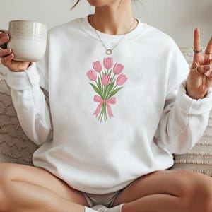 K&ouml;nnte beinhalten: Wei&szlig;es Sweatshirt mit einem gestickten Strau&szlig; rosa Tulpen, die mit einem rosa Band gebunden sind. Das florale Design ist auf der Vorderseite des lang&auml;rmeligen Oberteils zentriert. Das Sweatshirt ist aus weichem Material.