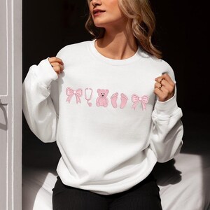 May include: White sweatshirt with pink embroidered designs, including bows, a stethoscope, a teddy bear, and baby footprints. The sweatshirt is a crew neck style.