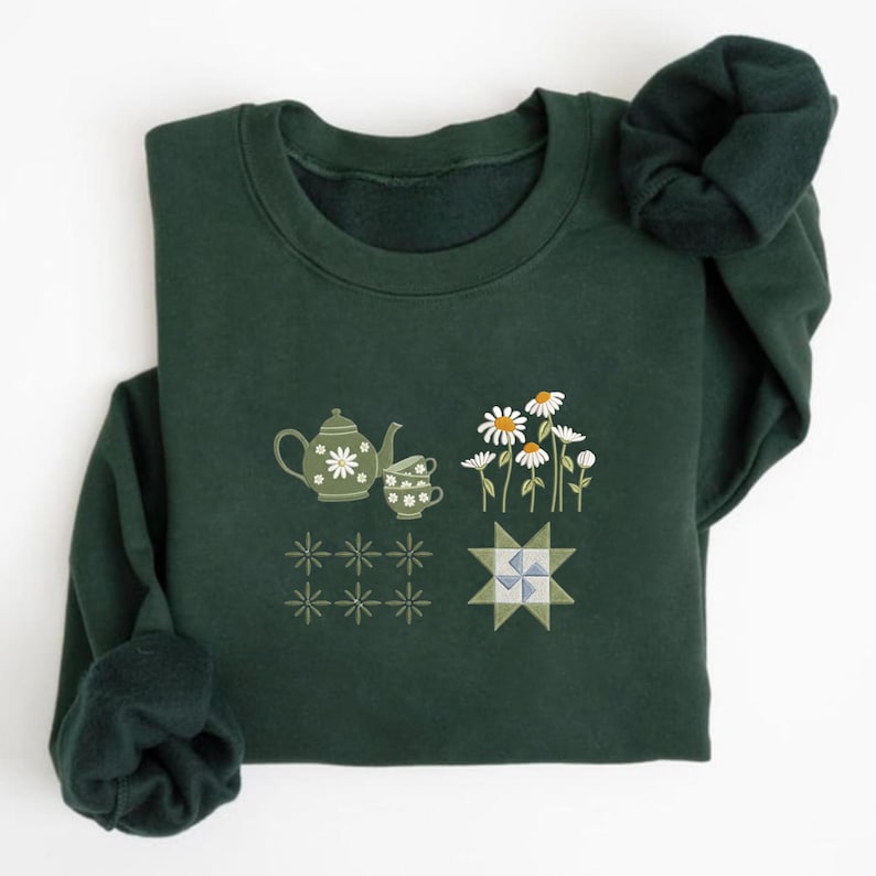 K&ouml;nnte beinhalten: Dunkelgr&uuml;nes Sweatshirt mit floralen und geometrischen Designs. Das Design umfasst eine Teekanne, eine Teetasse und G&auml;nsebl&uuml;mchen sowie Stern- und Quiltblockmuster. Das Sweatshirt hat einen Rundhalsausschnitt und lange &Auml;rmel.