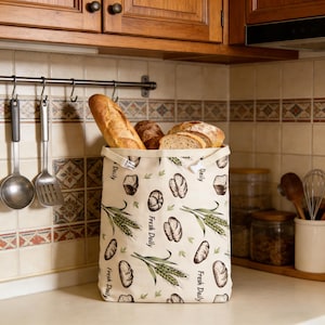 May include: A beige bread bag filled with various types of bread, including a baguette and sliced loaves. The bag features a repeating pattern of bread illustrations and the text "Fresh Daily". The bag is on a kitchen countertop.