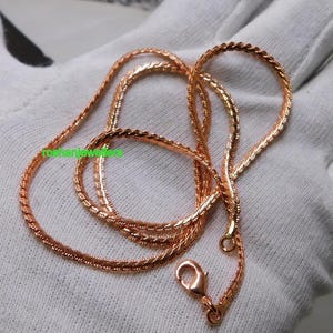 Op de afbeelding: Een roségouden ketting met een karabijnsluiting. De ketting heeft een gevlochten ontwerp en wordt gepresenteerd op een wit oppervlak. De ketting is een delicaat sieraad.