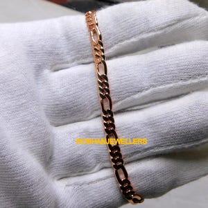 May include: A rose gold Figaro chain bracelet featuring a repeating pattern of three small circular links followed by one elongated oval link. The metal has a polished, reflective finish. The text ROSHANJEWELLERS is printed in yellow over the chain.