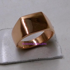 May include: A rose gold signet ring with a rectangular face. The ring has a polished finish and a smooth, rounded band. The ring is shown on a light grey surface. The ring is likely made of a precious metal.