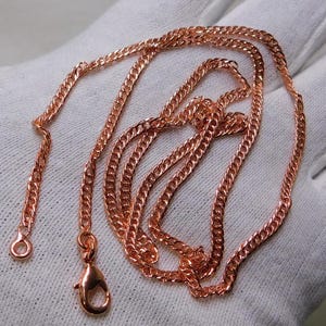 Peut inclure: Un collier chaîne gourmette en or rose avec un fermoir mousqueton et une petite boucle à l'extrémité. La chaîne est posée sur une surface blanche. La chaîne est composée de petits maillons imbriqués.