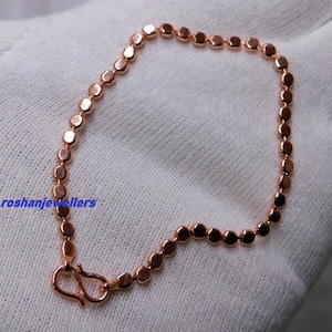 Puede incluir: Una pulsera de tono oro rosa compuesta por pequeñas cuentas metálicas redondeadas y uniformes unidas en una cadena continua. La pulsera presenta un sencillo cierre de gancho en forma de S en un extremo. El texto roshanjewellers es visible en azul.