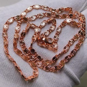 May include: A rose gold necklace with a delicate chain featuring a repeating floral design. The necklace is displayed on a white surface, showcasing its intricate details and warm metallic color. The clasp is visible, indicating the necklace is ready to wear.