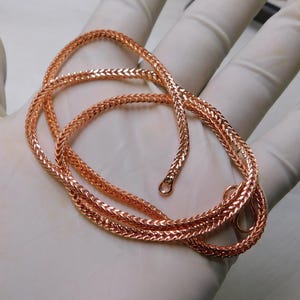 May include: A rose gold wheat chain necklace. The chain is made of interwoven links, creating a textured surface. The necklace is displayed on a light background, highlighting its warm metallic color and intricate design. The chain has a lobster clasp.