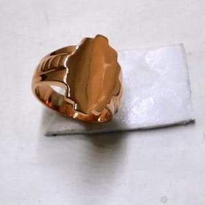 Pode incluir: Um anel dourado com uma grande face oval irregular. O anel tem uma banda grossa com cristas decorativas nas laterais. A face do anel é lisa e polida, refletindo a luz.