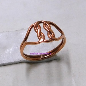 Puede incluir: Un anillo de color cobre con un diseño de nudo celta. El anillo presenta tres bucles entrelazados en el centro, con un acabado liso y pulido. El anillo está sobre una superficie de color claro. El texto "roshanjewellers" es visible.