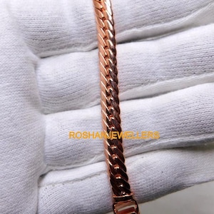 Peut inclure: Un bracelet de couleur cuivre métallique présentant un design en chaîne torsadée semblable à une corde avec un fermoir rectangulaire à l'extrémité. Le texte ROSHANJEWELLERS est imprimé en jaune sur le bracelet sur un fond en tissu blanc.