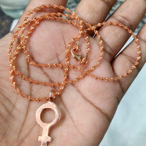 May include: A rose gold necklace featuring a pendant with the female symbol. The chain has a twisted rope design and a lobster clasp closure. The necklace is displayed on a hand.