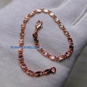Puede incluir: Una pulsera de cadena en oro rosa que presenta una serie de eslabones ovalados interconectados con un patrón texturizado de rayos de sol o estrellas en cada eslabón. La pulsera incluye un cierre de mosquetón y el texto ROSHANJEWELLERS es visible en azul.