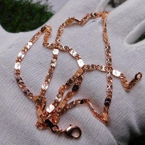 May include: A rose gold-toned chain necklace featuring flat, diamond-cut links that create a reflective, textured surface. The chain includes a secure lobster claw clasp at one end, showcasing a metallic finish with a warm, reddish-pink hue.