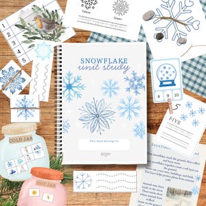 Puede incluir: Un cuaderno encuadernado en espiral titulado "Snowflake Unit Study" con ilustraciones de copos de nieve azules. Los materiales educativos incluyen tarjetas de números, ejercicios de trazado y actividades "Cold Jar" y "Hot Jar", todo sobre una superficie de madera.