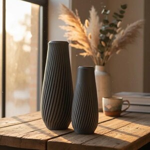 Puede incluir: Dos jarrones grises oscuros texturizados con un diseño en espiral sobre una mesa de madera. El jarrón más alto mide unos 30 cm de alto, y el más bajo, unos 20 cm. La mesa está cerca de una ventana soleada.