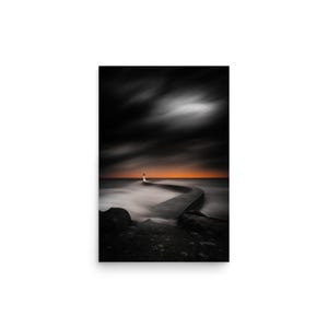 May include: A black and white photograph of a lighthouse at the end of a curved pier. The sky is dark with streaks of clouds, and the horizon glows orange. The water appears blurred, creating a sense of movement.