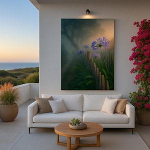 May include: A large canvas art print depicting a serene garden scene with purple flowers, a wooden fence, and a misty background. The artwork hangs above a white sofa with neutral-toned pillows on an outdoor patio. A round wooden coffee table and potted plants complete the setting.