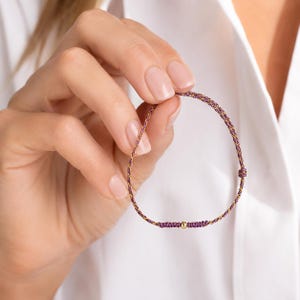May include: A woven bracelet with purple and gold threads, featuring a small gold bead. The bracelet is held against a white shirt, showcasing its delicate design and colour combination. The bracelet is a simple yet elegant accessory.