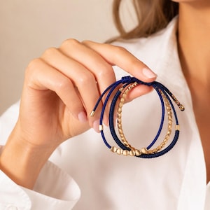 Peut inclure: Trois bracelets sont présentés, combinant des cordons bleu marine et des détails en métal doré. Un bracelet a un design de chaîne, tandis que les autres ont un design de cordon avec des perles dorées. Les bracelets sont tenus contre une chemise blanche.