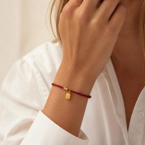 Pode incluir: Uma pulseira trançada vermelha com um pequeno berloque dourado e um fecho dourado. O berloque é retangular com um design detalhado. A pulseira é usada em um pulso.