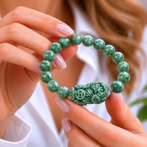 Può includere: Un braccialetto di giada verde con perline rotonde e un elemento centrale scolpito. Il braccialetto è mostrato, mettendo in risalto la consistenza lucida e il design dettagliato dell'elemento centrale.