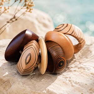 Peut inclure: Plusieurs bracelets en bois de différentes formes et couleurs. Certains sont brun foncé, d'autres sont brun clair avec des motifs rayés noirs. Les bracelets sont posés sur une surface en pierre claire, avec un arrière-plan flou de l'océan.