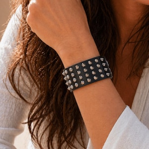 Puede incluir: Brazalete de cuero negro con tachuelas cónicas plateadas. La pulsera ancha tiene tres filas de tachuelas. El accesorio se lleva en la muñeca de una persona. Un accesorio atrevido.