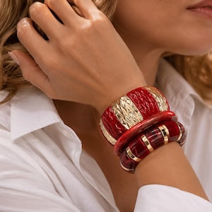 Può includere: Un polso adornato da tre braccialetti impilati. Il braccialetto superiore è rosso con dettagli dorati e un design strutturato. Gli altri due braccialetti sono rossi con dettagli dorati. La persona indossa una camicia bianca abbottonata.
