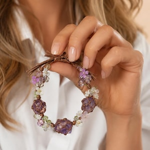 Peut inclure: Un bracelet artisanal orné de grosses pierres d'améthyste brutes, entrecoupées de petites fleurs délicates dans les tons de violet, rose et blanc. Le bracelet est maintenu par un cordon marron et est présenté sur une chemise blanche.
