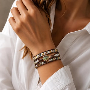 Peut inclure: Bracelet enroulé avec plusieurs rangées de perles colorées vertes, blanches et marron. Le bracelet a une bande en cuir marron et une pierre verte en forme de goutte. Le bracelet est porté sur un poignet.