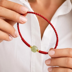 Peut inclure: Un bracelet tressé rouge avec une perle carrée vert clair et des accents dorés. Le bracelet est tenu contre une chemise blanche, mettant en valeur le design et le contraste de couleurs du bijou. C'est un bijou délicat.
