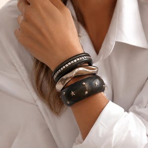 May include: A collection of bracelets in various styles. The bracelets include a black leather band with silver studs, a silver cuff, and a dark wooden bracelet with star-shaped studs. The bracelets are stacked on a wrist.