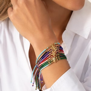 Könnte beinhalten: Eine Sammlung farbenfroher Schnurarmbänder mit goldfarbenen Ankh-Anhängern. Die Armbänder sind in verschiedenen Farben erhältlich, darunter Blau, Grün, Rot und Schwarz. Die Armbänder sind mit verstellbaren Knoten gebunden.