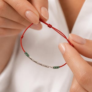 Peut inclure: Bracelet en ficelle rouge avec des perles cylindriques argentées et des nœuds verts. Le bracelet est tenu entre les mains, mettant en valeur le design et le contraste des couleurs. Un bijou simple et élégant.