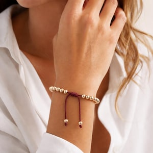 Peut inclure: Bracelet de perles dorées avec un cordon bordeaux et une fermeture réglable. Le bracelet est composé de petites perles dorées rondes enfilées ensemble, créant un accessoire délicat et élégant. Le cordon bordeaux ajoute une touche de couleur.