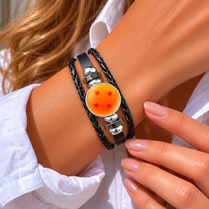 Könnte beinhalten: Schwarzes Lederarmband mit einem runden, orangefarbenen Anhänger mit vier roten Sternen. Das Armband hat silberfarbene Metallakzente und ein geflochtenes Lederband. Der Anhänger ist der Mittelpunkt des Armbands.