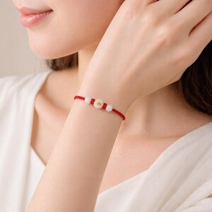 Op de afbeelding: Rode draadarmband met witte kralen en een goudkleurige bedel. De armband wordt om een pols gedragen, met de arm en een deel van het gezicht van de persoon zichtbaar. De armband heeft een eenvoudig ontwerp, waarschijnlijk voor dagelijks gebruik.