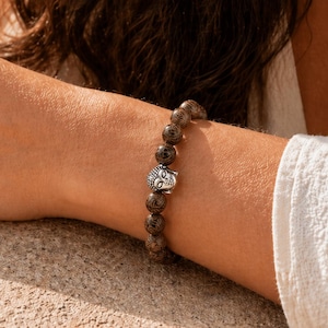 May include: A beaded bracelet featuring dark brown, patterned beads and a silver-colored Buddha head charm. The bracelet is worn on a wrist, showcasing the jewelry's design and texture. The beads appear to be approximately 0.5 inches in diameter.