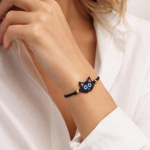 Op de afbeelding: Een zwart kralenarmband met een kattengezichtontwerp in roze en blauw. Het kattengezicht is gemaakt van kleine kralen en de armband heeft een zwart koord met goudkleurige kralen. De armband wordt om een pols gedragen.