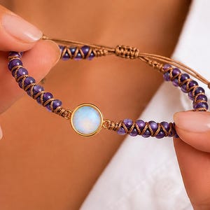 Peut inclure: Un bracelet fait à la main avec une pierre centrale opalescente ronde sertie d'or. Le bracelet est composé de petites perles violettes et d'un cordon marron, créant un design délicat. Le cordon réglable permet un ajustement confortable.