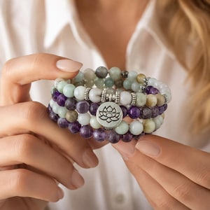 May include: A collection of beaded bracelets in shades of purple, green, white, and grey. The bracelets are held in a person's hands. A silver charm with a lotus flower design hangs from one bracelet. The beads appear to be semi-precious stones.