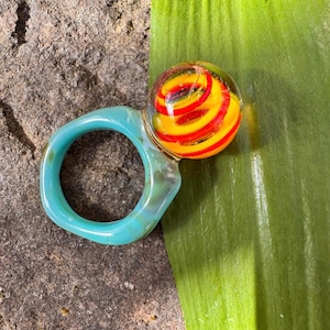 May include: A distinctive ring with a clear glass orb containing a red and yellow spiral design. The ring's band is a light teal colour with a marbled effect. The ring is set on a textured stone surface.
