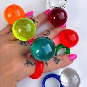 May include: A collection of seven chunky rings featuring large, smooth, spherical tops in vibrant translucent colors including orange, lime green, teal, red, dark red, clear, and a blue ring with a clear sphere, all made of polished resin.