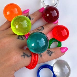 Op de afbeelding: Een collectie grove ringen met grote, bolvormige, doorschijnende harsknoppen in levendige kleuren zoals oranje, limoengroen, donkerrood, taling, felrood en transparant, elk bevestigd aan een bijpassende of contrasterende ronde ringband.