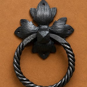 Può includere: Un battente per porta in metallo nero caratterizzato da un anello circolare a corda intrecciata fissato a una piastra decorativa a forma di fiore stilizzato con cinque petali. La piastra presenta tre piccoli fori per il montaggio.