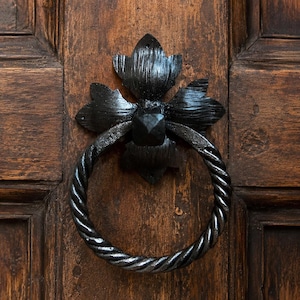 Puede incluir: Primer plano de un aldaba de metal negro con un asa de anillo retorcido y una placa trasera floral decorativa. La aldaba está montada en una puerta de madera marrón oscura con vetas visibles.