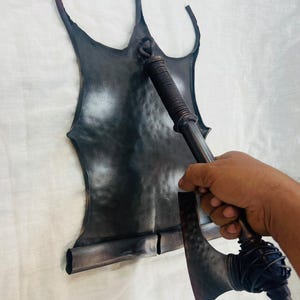 Puede incluir: Un escudo de metal oscuro martillado con una parte superior puntiaguda y un borde inferior enrollado, junto con un hacha de metal oscuro a juego con una hoja martillada texturizada y un mango envuelto, ambos con un acabado industrial desgastado.