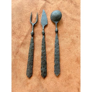 May include: Three-piece rustic forged iron flatware set. The set includes a fork, a knife, and a spoon. The utensils have textured handles and are displayed on a brown surface. The fork has two tines, the knife has a triangular blade, and the spoon has a round bowl.