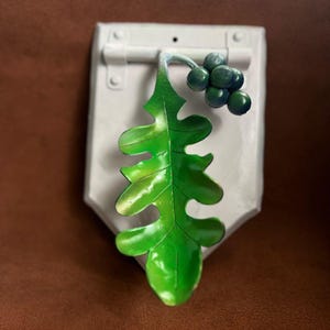 May include: A white, shield-shaped wall decoration featuring a green leaf and dark green berries. The leaf has detailed veining and a glossy finish. The berries are clustered. The piece has a rustic, handcrafted appearance.