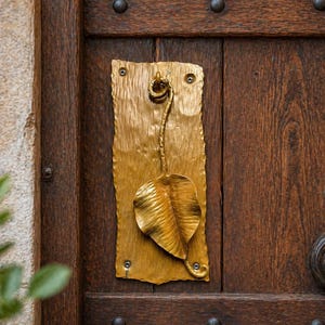 Könnte beinhalten: Ein goldfarbener Türklopfer in Blattform, montiert auf einer strukturierten rechteckigen Platte. Die Platte ist an einer dunkelbraunen Holztür mit Metallstiften befestigt. Das Blattdesign hat detaillierte Adern.
